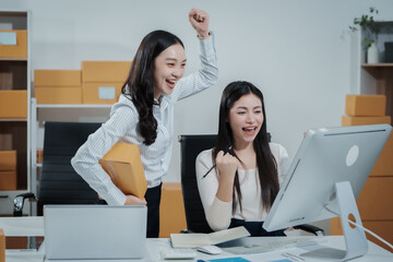 Two Asian female entrepreneurs celebrate a major online sales achievement,cheering happily while checking their laptop in an e-commerce office surrounded shipping boxes,excitement,business success