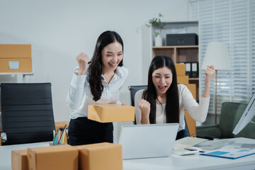 Two Asian female entrepreneurs celebrate a major online sales achievement,cheering happily while checking their laptop in an e-commerce office surrounded shipping boxes,excitement,business success