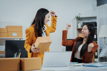 Two Asian female entrepreneurs celebrate a major online sales achievement,cheering happily while checking their laptop in an e-commerce office surrounded shipping boxes,excitement,business success