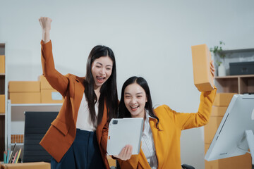 Two Asian female entrepreneurs celebrate a major online sales achievement,cheering happily while checking their laptop in an e-commerce office surrounded shipping boxes,excitement,business success