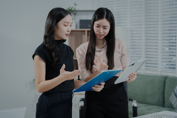 Two Asian businesswomen working together in office, holding clipboard and documents, discussing...