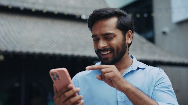 Portrait of Indian man uses sign language on video call on city street. Smiling bearded male gestures talking on smartphone in urban district. Non verbal communication method