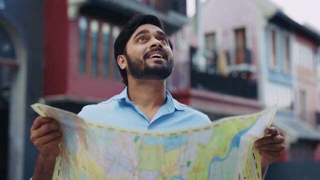 Portrait of bearded Indian man reads map visiting foreign city on holiday. Smiling South Asian traveler seeks famous sightseeing with paper plan in urban block. Travel destination
