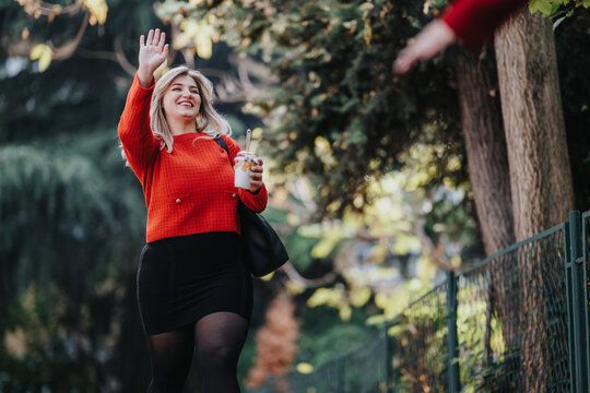 A cheerful woman in a bright red knit sweater smiles and waves as she walks along a tree-lined path, carrying a black bag and a jar, enjoying a casual outdoor moment.