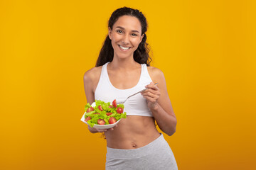 Cheerful fit woman holds a bowl of fresh vegetable salad and fork, smiling brightly. The vibrant yellow-orange wall enhances the healthy dieting theme and her positive energy.