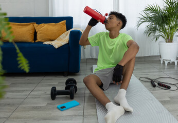 Latino teenager sitting and resting on a mat in his living room while drinking water from a red bottle, illustrating hydration and a healthy lifestyle