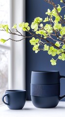 Two navy blue ceramic mugs sit on a white surface next to a decorative branch with bright green leaves, with a window and dark blue wall in the background.