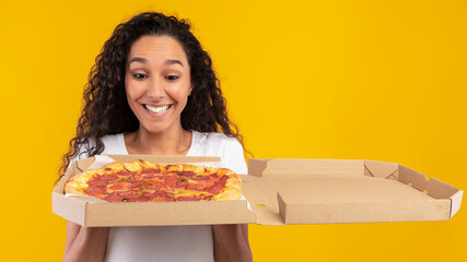 A joyful young lady gazes at an open pizza box filled with tasty Italian pizza. She stands in front of a bright yellow-orange wall, celebrating her food delivery.