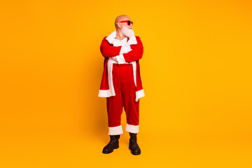 Santa in a red fur coat with white trim and boots stands with arms crossed against a bright yellow background Christmas themed