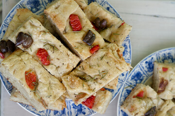 Focaccia squares piled on a plate, featuring olives, rosemary and cherry tomatoes. Flatten top view