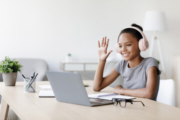 A young afro american pupil wearing headphones smiles while watching a video lesson on her laptop in a home setting. She waves her hand, enjoying remote learning during social distancing.