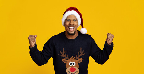 Obraz premium A man with an enthusiastic expression wears a festive reindeer sweater and a Santa hat. He raises his fists in celebration against a bright yellow background, embodying holiday cheer.
