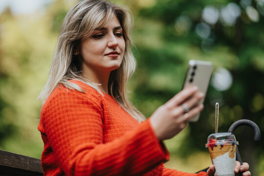 A woman in a bright orange sweater sits on a park bench, holding a smartphone for a selfie while enjoying an iced coffee. Lush greenery in the background conveys a relaxed outdoor moment. - Powered by Adobe