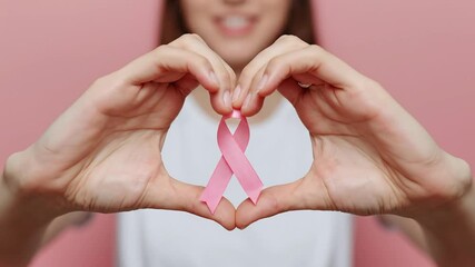 Hands forming heart shape around pink ribbon, symbolizing breast cancer awareness, support, and hope on a soft pink background.