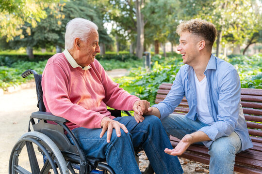 Smiling grandson holding grandfather&rsquo;s hand beside grandfather seated in wheelchair on park bench