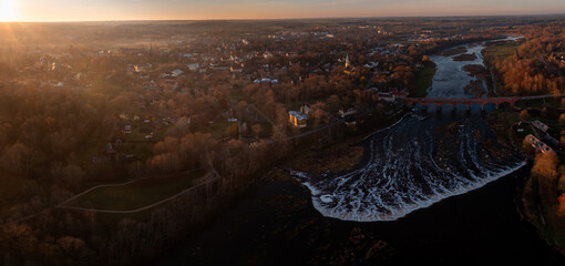 Aerial view shows Venta Rapid and the 19th century red brick bridge in Kuldiga at sunset, wide waterfall in foreground, warm light on church spires and streets.