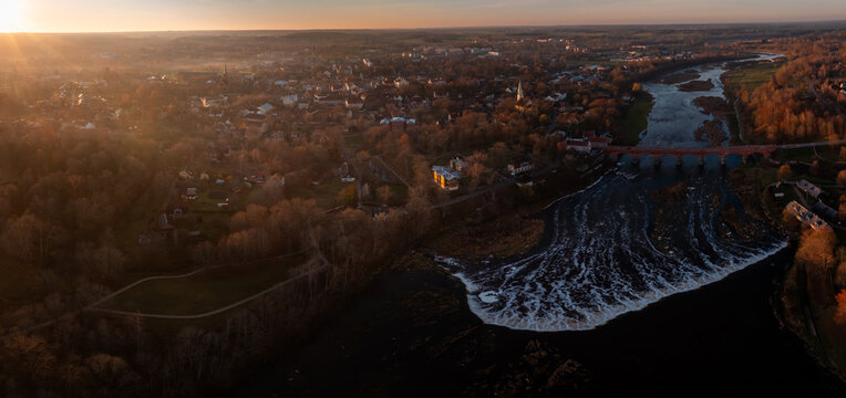 Aerial view shows Venta Rapid and the 19th century red brick bridge in Kuldiga at sunset, wide waterfall in foreground, warm light on church spires and streets.