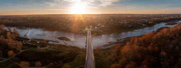 Aerial view shows Venta Bridge leading to Kuldiga at sunset, warm light on water channels and autumn trees, church spires in distance, centered road and flare.