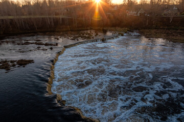 Venta Rapid in Kuldiga Latvia shows wide stone ledge and frothy flow at golden hour. Low sun flares over leafless trees and houses, late autumn or early winter.