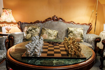 A vintage sitting room frames a green marble table with silver and gold chessmen. Warm lamplight glows on carved wood and brocade as an evening match is about to begin.