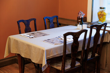 A corner dining table near a window sits set with menus, cutlery, salt and pepper, and pink flowers. A large jar of lemons rests on the sill in daytime light.