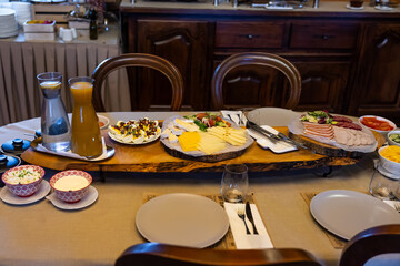 A long live edge board holds sliced cheeses, fanned cold cuts, boiled eggs, and bowls of salads and spreads, with carafes of water and orange juice on a set table.