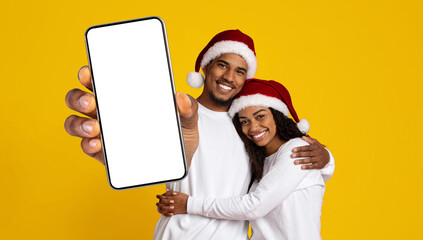 A joyful couple poses together with an empty smartphone screen against a bright yellow background. They wear matching Santa hats and have cheerful expressions, celebrating the holiday spirit.