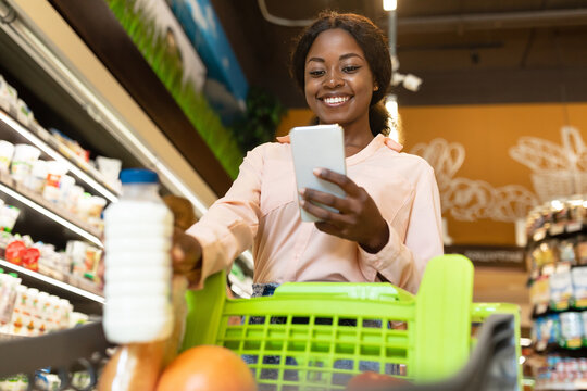 A young woman smiles at her smartphone while pushing a shopping cart in a colorful grocery store filled with fresh produce and dairy products. - Powered by Adobe