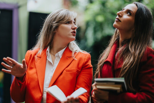 Two women stand outdoors, one in an orange blazer and the other in a red coat, engaging in a animated discussion while holding books. Modern, fashionable, and collaborative vibe.