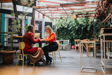 Two women sit across from each other at a colorful outdoor cafe, talking over a table.