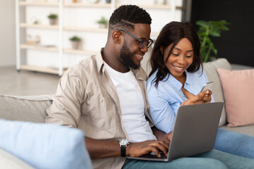 A cheerful black couple relaxes on their couch in the living room, engaging with a cellphone and a laptop. They share a moment of joy while using their devices, showcasing modern lifestyle.