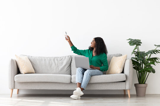 A young woman with curly hair sits comfortably on a light gray couch, smiling as she uses her smartphone. The bright room features a potted plant and soft cushions. - Powered by Adobe
