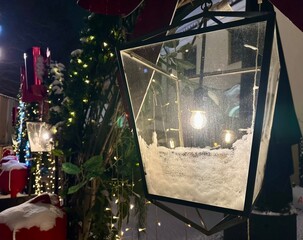 Decorative large lantern with snow inside, illuminated by warm light, surrounded by festive holiday decorations, creating a cozy winter atmosphere with twinkling lights