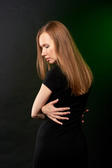 Naklejka premium A beautiful young woman in a black dress stands in front of the camera on a dark background during a photo shoot in the studio