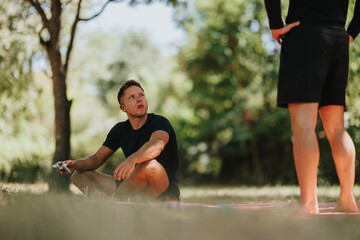 Two men outdoors in a sunny park scene: one sits on the grass in a black shirt while the other stands nearby, suggesting a casual workout break and friendly chat.