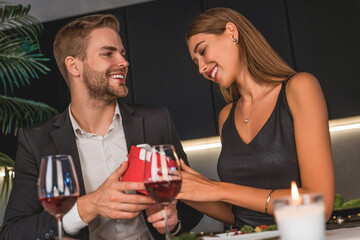 Young romantic couple sitting at home kitchen and man giving gift box his female during romantic dinner. Present sharing on date. Bonding and relationship