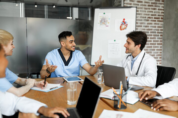 Cheerful multiracial doctors men and women in uniforms sitting aroung table at clinic, having conversation, using modern gadgets, sharing professional experience, medical conference concept