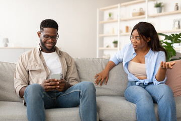 A couple sits on a couch in a living room. The woman looks angry and is gesturing while the man is focused on his phone, seemingly ignoring her concerns during a tense moment.