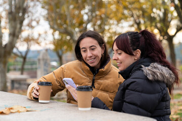 Two smiling women friends sharing a moment outdoors, looking at a smartphone and drinking coffee on a fall day