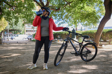 Woman in sportswear drinking water while standing next to a mountain bike in a sunny park.