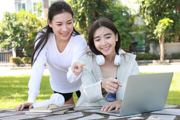 Pretty cheerful asian woman in casual clothes browses laptop computer connected to 4g internet updates software uses modern technologies poses outdoor looks gladfully away with her friend.