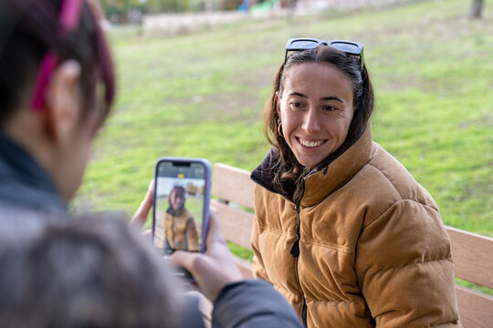 Woman smiling for a friend taking a smartphone portrait in a park, enjoying social media and modern communication