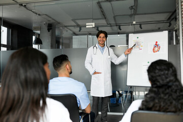 Cheerful young man chief doctor standing by presentation board, having speech, multiracial team of professional doctors attending medical conference or training, listening to speaker