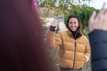 Young woman waving hello or goodbye with a friendly smile, greeting or seeing someone off outdoors in autumn
