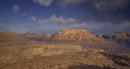 The rugged peaks of a desert plateau rise against a dramatic sky filled with clouds as the sun sets, casting shadows across the arid terrain and highlighting the natural beauty of the landscape.