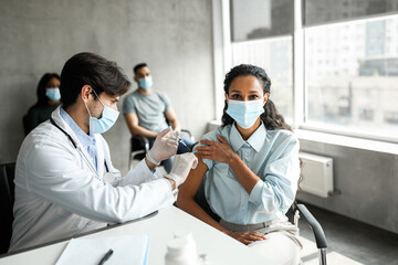 Young woman patient getting intramuscular shot against coronavirus at walk-in vaccination site, male doctor making injection in shoulder for lady, wearing protective face masks, copy space