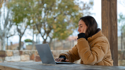 Woman working on laptop outdoors, sitting on a wooden picnic table, managing her remote work...