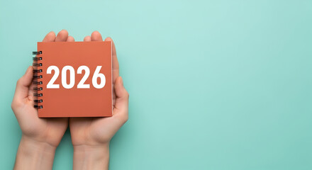 A pair of hands gently holds a small, square, coral-colored notebook with the year 2026 emblazoned in white, against a serene, light blue background.