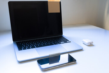 Laptop with covered webcam, reflective smartphone and wireless earbuds on white table; soft daylight and copy space suggest focus, privacy, study habits and teen tech lifestyle.