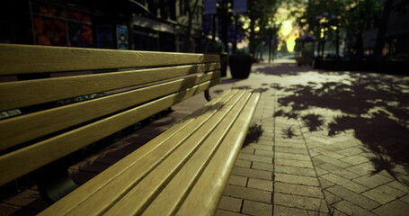 Dappled sunlight casts intricate shadows on a wooden park bench in a bustling city park during the early evening. Trees and buildings create a lively atmosphere.